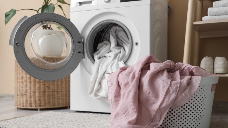 A laundry room with a basket full of sheets and an open machine filled with sheets