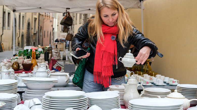 Blonde woman in red scarf and black jacket lifts tea pot from array of plates at a flea market