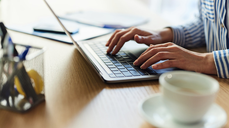 A woman's hands are seen typing on a laptop next to a coffee cup