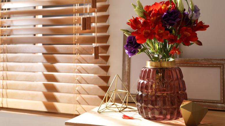 Wooden blinds in the window next to a table with red and violet flowers in an amethyst colored vase