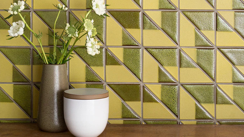 A vase of flowers and a small jar sit on a wooden countertop in front of a mustard and green tile backsplash