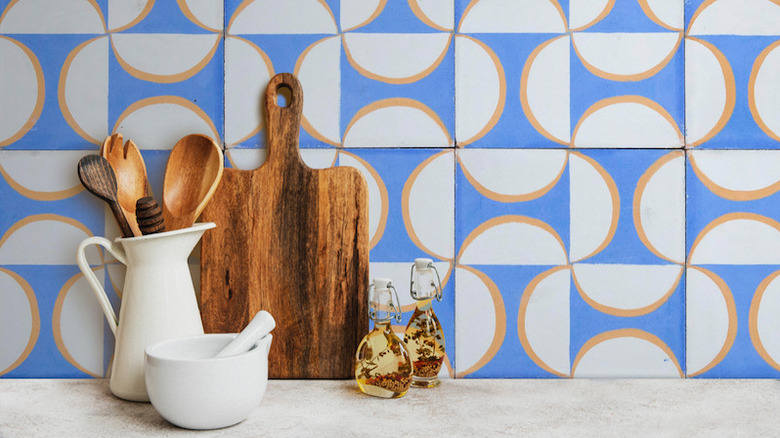 A cutting board and pitcher full of cooking utensils sits on a white countertop in front of a colorful mosaic backsplash