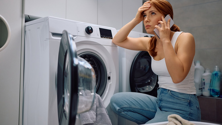 A stressed woman on the phone with washer door open and clothes spilling out