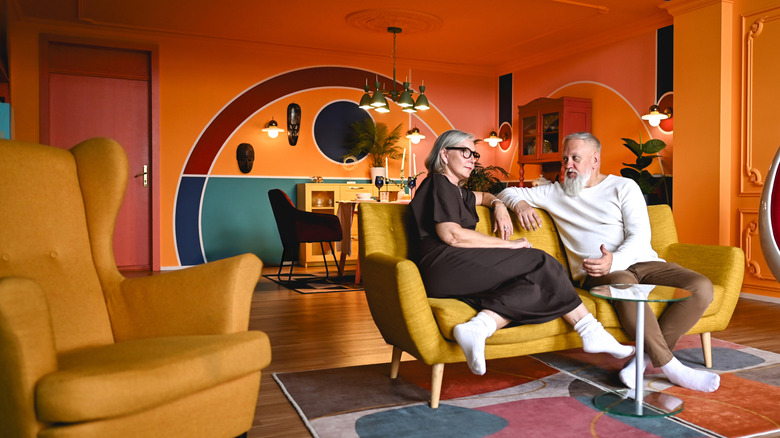 Man and woman on sofa in funky modern room with orange walls, yellow chairs, and geometric carpet and accents
