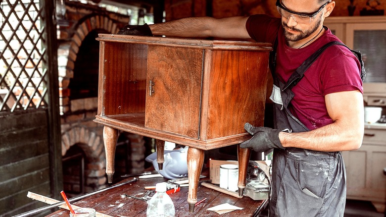 Person wearing protective eye wear carries a small antique cabinet to a work surface to restore