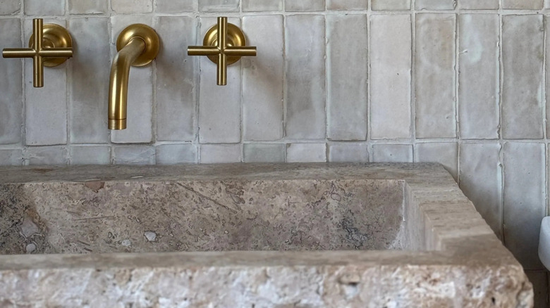A limestone sink with a white zellige tile backsplash and soft gold faucet.