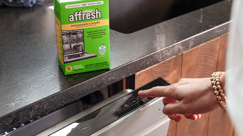 A hand turning on a dishwasher with a box of Affresh dishwasher cleaning tablets on the counter above