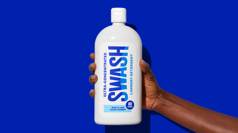 A hand holding a bottle of Swash laundry detergent against a blue background