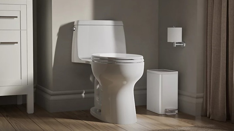 A small white trash can tucked in beside a toilet