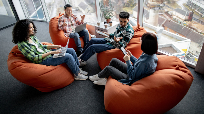 A diverse group of young professionals has a work meeting while sitting in orange bean bag chairs.