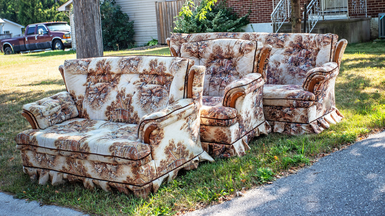 A 1970s couch, love seat, and arm chair in a matching autumnal print are sitting by the side of the road in front of a home.