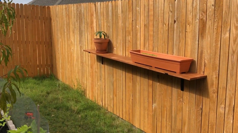 A simple shelf made from scrap wood attached to a wood fence with potted plants on top