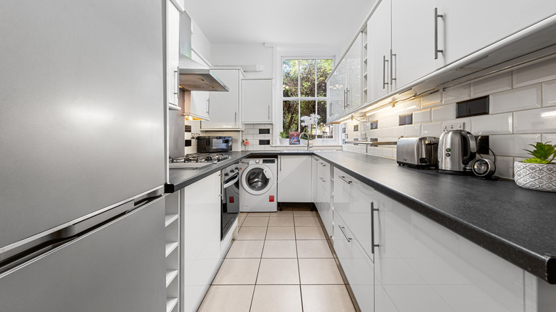 Galley kitchen with white cupboards and appliances and a window at the far end
