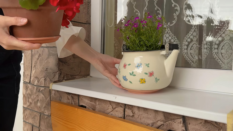 A hand placing a teapot with flowers inside it on a windowsill