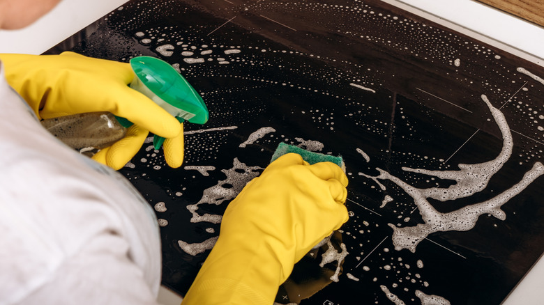 Close-up of a person cleaning a glass stove with a bottle of cleaner in one hand