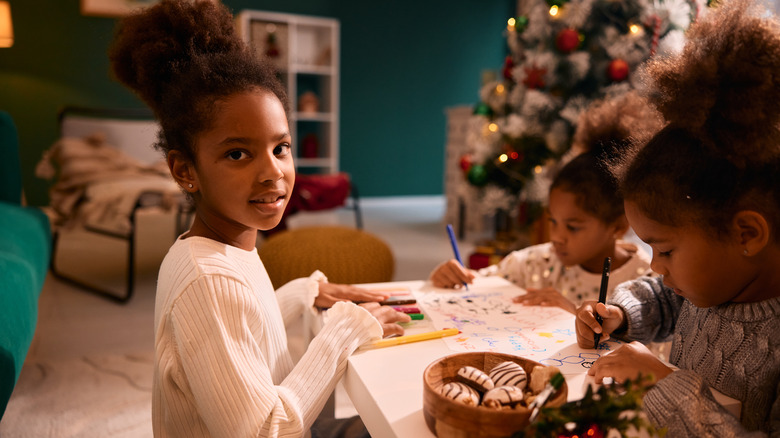 Three siblings happily color together at a kids' table by a Christmas tree