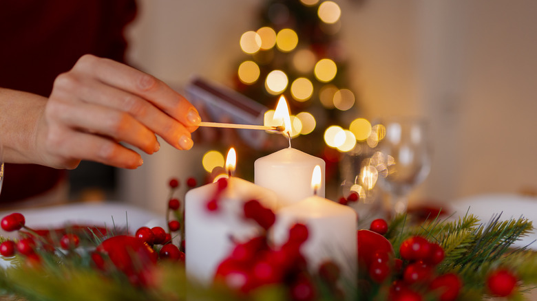 Hands light a white candle surrounded by holiday greenery