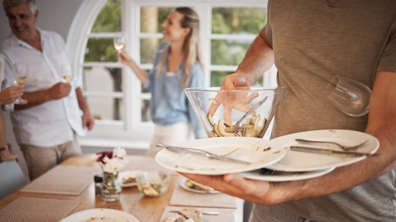 A man clears several plates at once as guests mingle behind him