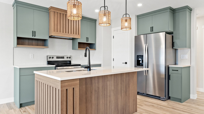 A kitchen with light green cabinets, stainless steel refrigerator, and a white island with a wood base
