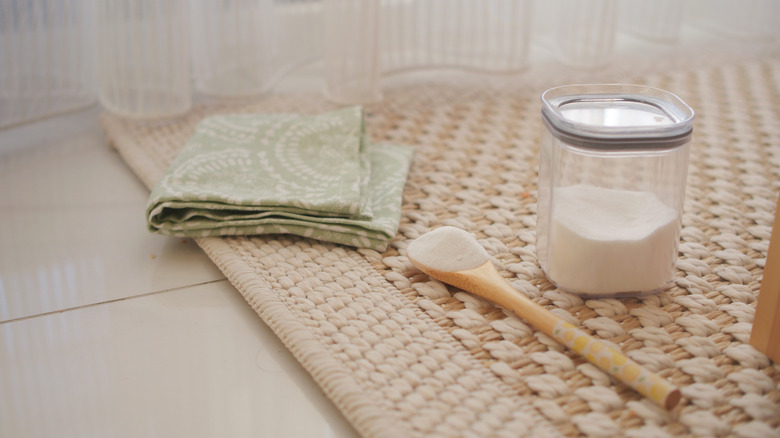 A baking soda jar, spoon with some baking soda on it, and cloth on natural fiber carpet