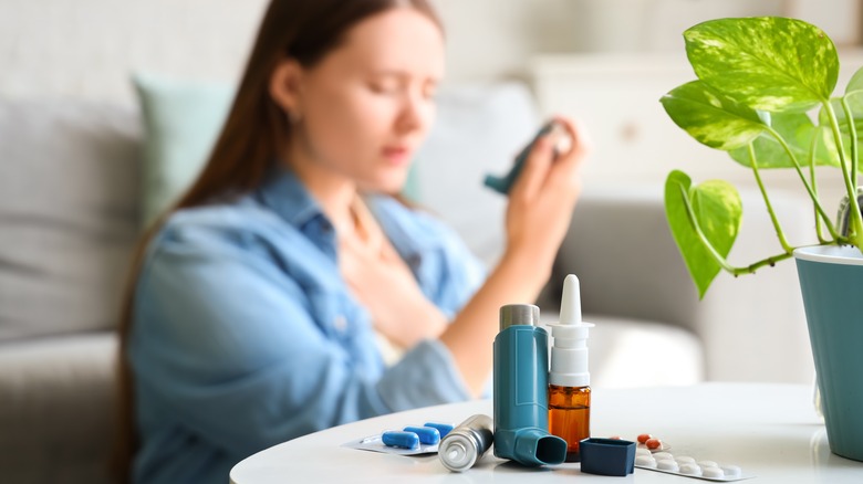 A woman using an inhaler next to a coffee table with allergy medicine.