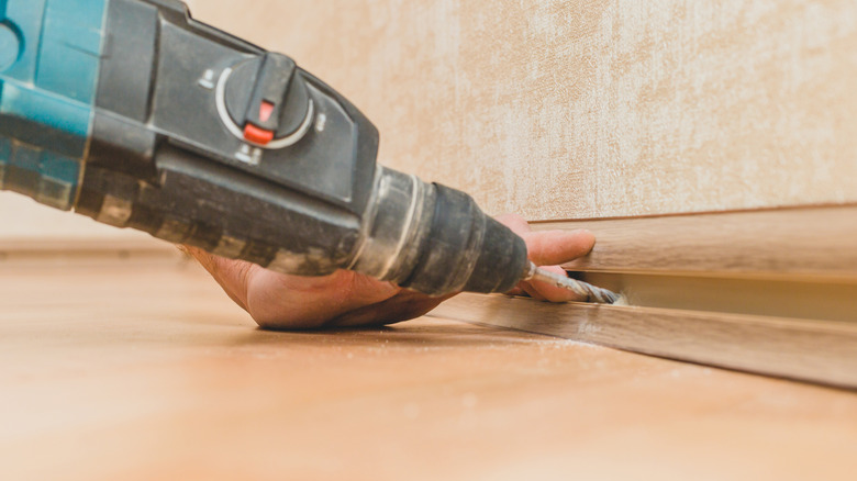 A hand drilling a hotle into the baseboard of a wall.