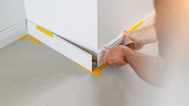 A pair of hands installing a white trim baseboard on a wall.