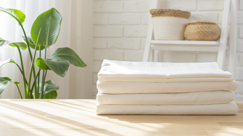 Folded white sheets on a wooden table in a bright room with a plant and decor in the background