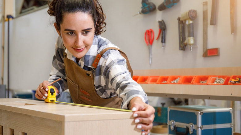 A person in flannel and overalls measuring a piece of furniture