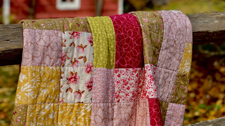 A quilt made up of various patterns in pink, white, yellow, and green hanging over an old wooden fence with a barn and fall leaves in the background