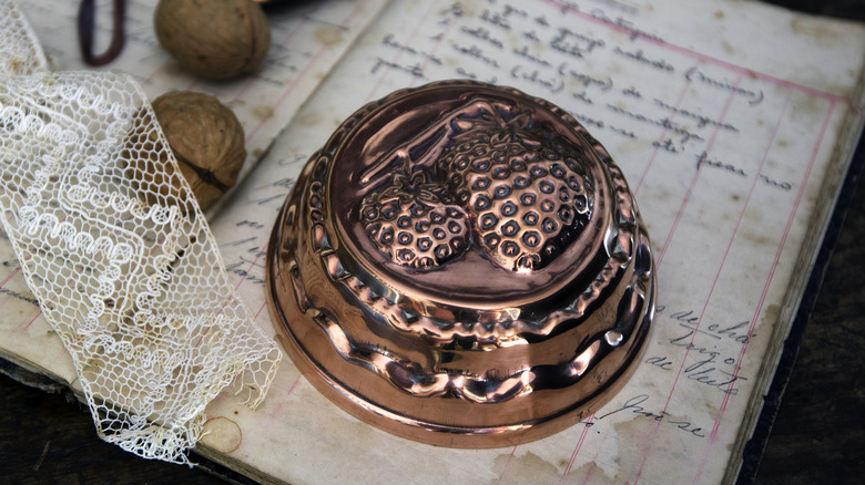 A copper cake mold with strawberries on top of a vintage book with nuts and lace on the side