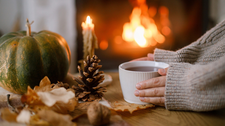 Two hands holding a white cup with a fireplace in the background and fall decor