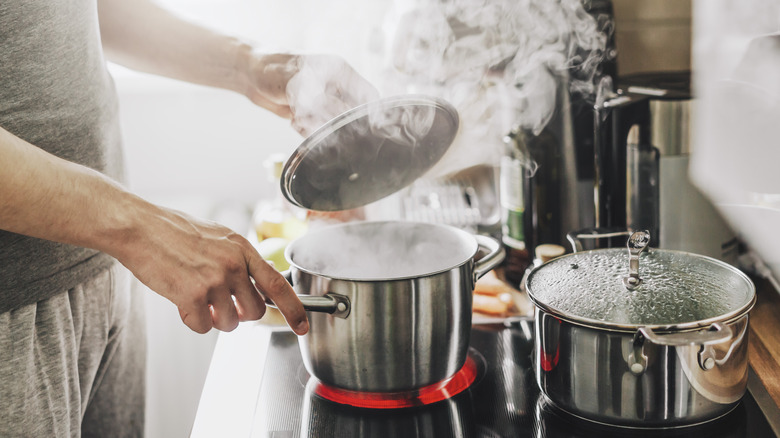 Young man cooking fresh food at home and opening lid of steaming pot