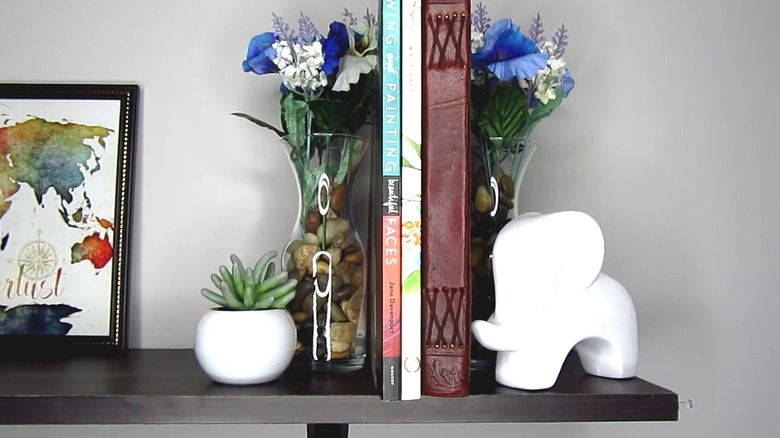 Two vases filled with pebbles and silk flowers used as bookends on a shelf with other decor pieces