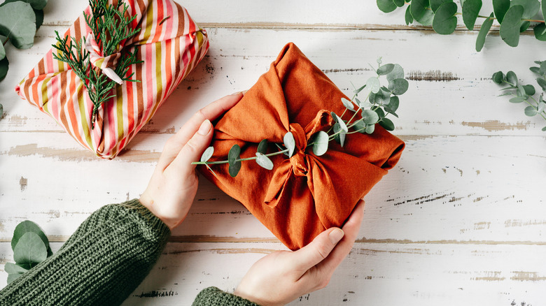 Person holding out a gift wrapped in fabric with eucalyptus tucked in it next to another fabric-wrapped present