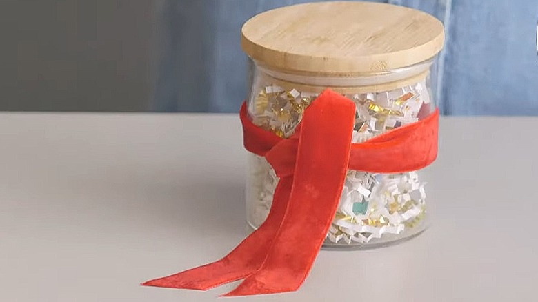 Small gift inside of a glass jar with shredded paper around it and a ribbon tied on the outside