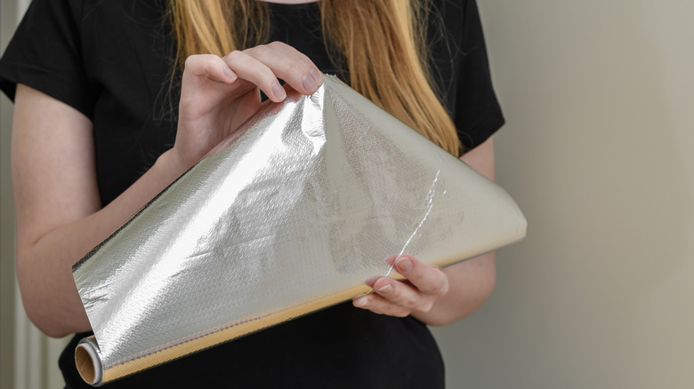 A woman pulls some aluminum foil from a roll she holds in her other hand.