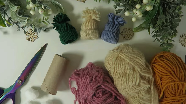 miniature yarn hats with a toilet paper roll, scissors, yarn, and winter decorations on a table