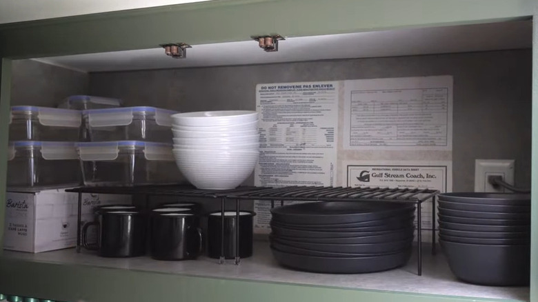 Black shelf riser in high kitchen cabinet with various dishes and mugs