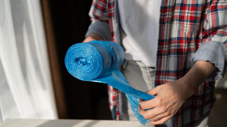 Person ripping a blue plastic bag off a roll, they wear a plaid shirt and denim jeans
