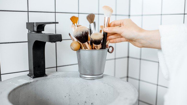 A hand takes a makeup brush from a small bucket on a bathroom sink.