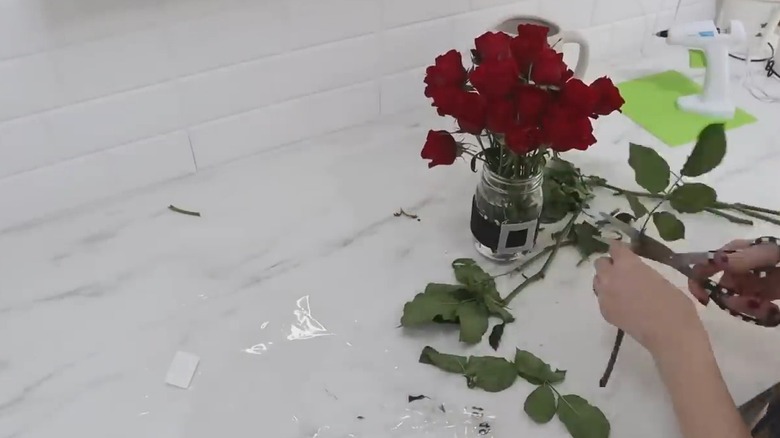 Red roses in a mason jar with a crafted Santa belt and someone cutting stems nearby