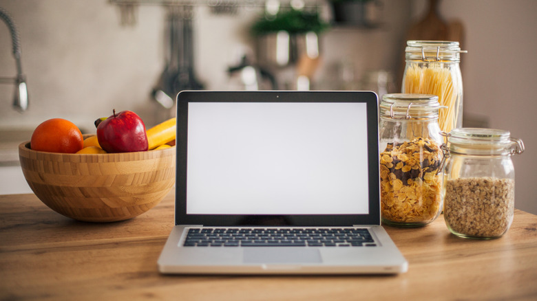 A laptop on a kitchen counter with jars of cereal to the side