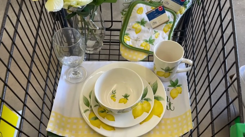 A shopping cart filled with lemon print kitchen linens, plates, and a matching mug from Dollar Tree.