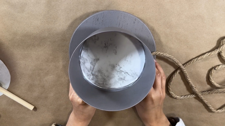 Hands attaching twine to the a painted trifle bowl on a wood base over a brown paper surface