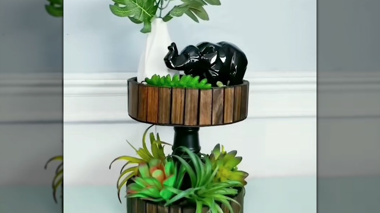 two-tiered wooden plant shelf with succulents, a white vase with greenery, and a black elephant layer on it