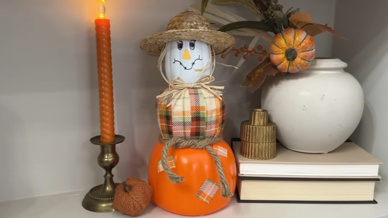 Scarecrow decoration made from three stacking pumpkins sitting by a candle and other decor