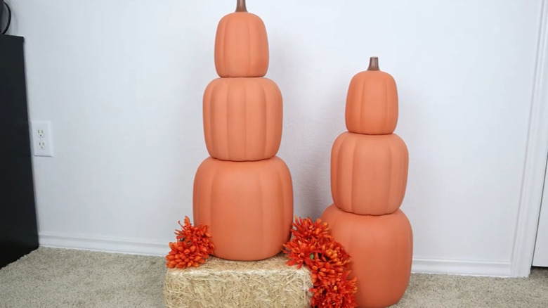 Two stacks of realistically painted pumpkins with one sitting on a hay bale and fall flowers as accents