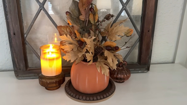 Single plastic pumpkin with fall foliage sticking out the top with a candle and old window behind it
