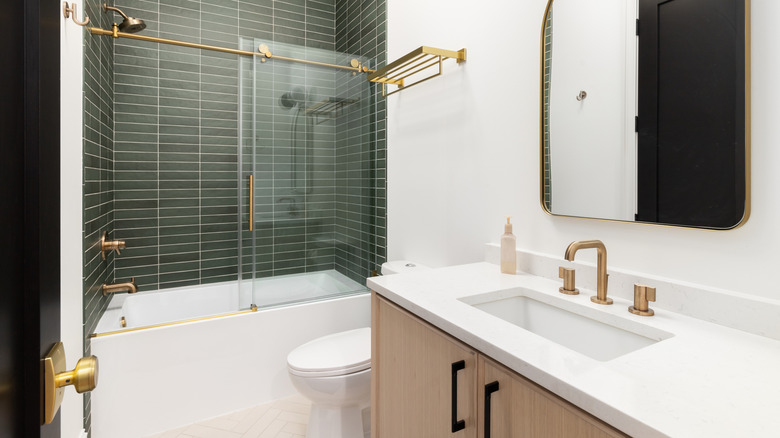 A bathroom with a wood vanity cabinet, gold hardware and faucet, and a shower with green subway tiles